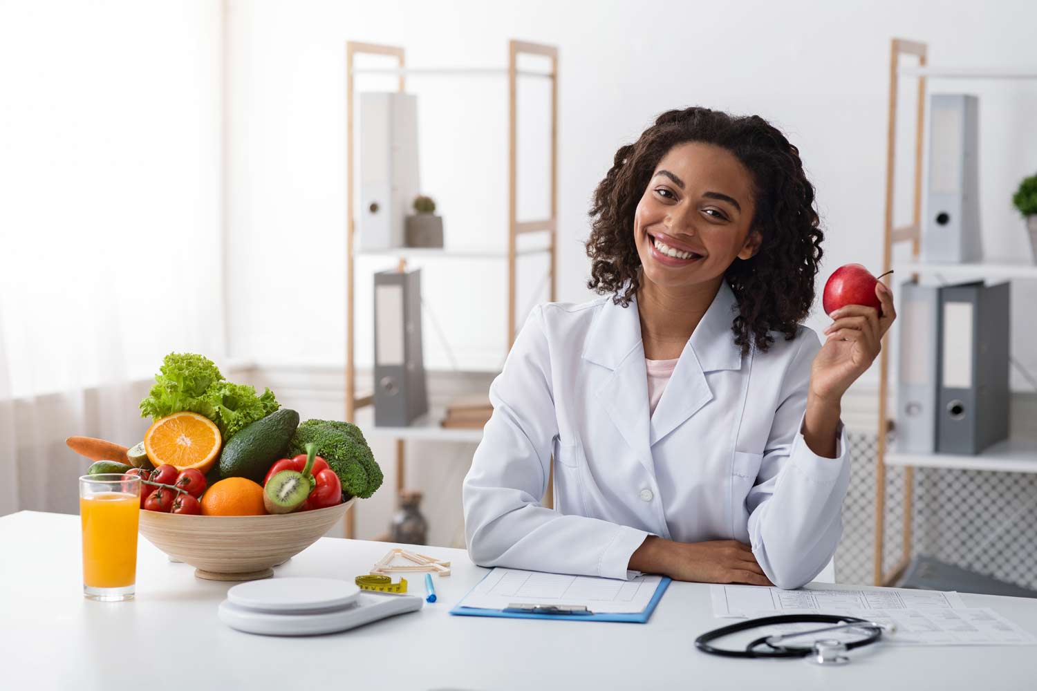 מדענית או רופאה בחלוק לבן ליד שולחן ועליו קערה עם פירות וירקות