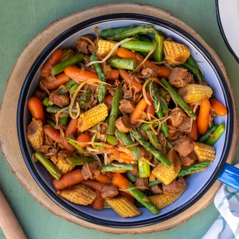 מבט מלמעלה על תבשיל עם פתיתי סויה, אספרגוס, גזר, תירס ונבטים בתוך מחבת לצד קערת אורז וכף הגשה