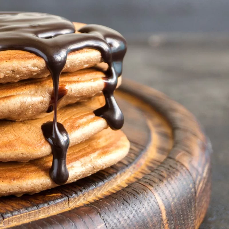 ערימה של פנקייק טבעוני על מגש עץ עם רוטב שוקולד מומס מעל